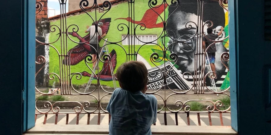 Irmão caçula de Rocha observa à janela enquanto ele e Hagah desenvolvem o mural. (Foto: Amanda Travassos)
